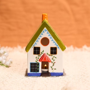 May include: A ceramic house-shaped candle holder with a lit candle inside. The house has a white body, green roof, and blue and red accents. The windows are black, and the chimney is orange. Decorative vines with red berries adorn the front.