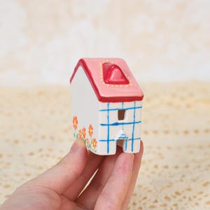 May include: A small, white ceramic house with a pink roof and red trim. The house features blue grid lines, a small window, and orange flower accents. The roof has a red chimney and is decorated with yellow dots.
