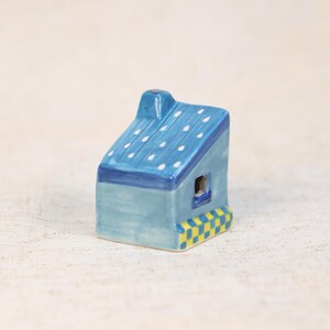 May include: A small, ceramic house figurine. The house has a light blue body, a dark blue roof with white polka dots, and a yellow and blue checkered base. A small window is visible on the side.