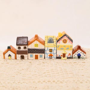 May include: A collection of six ceramic miniature houses in various colors and designs. Each house has unique architectural details, including different roof styles, window shapes, and door colors. The houses are arranged in a row, creating a charming village scene.