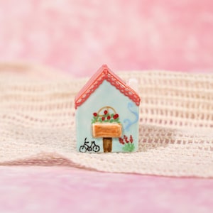 May include: A small ceramic house with a light blue facade, red roof, and floral decorations. The house has a bicycle and a flower box. The background is a pink surface with a white knitted texture.