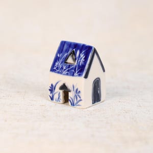 May include: A small ceramic house with a blue roof and white walls. The roof is decorated with a floral pattern. The house has a small triangular window and a black door. The base of the house has a floral design.