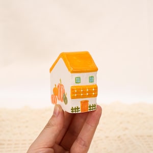 Könnte beinhalten: Ein kleines, handbemaltes Keramikhaus mit orangefarbenem Dach und Kürbisillustrationen. Das weiße Haus hat eine kleine Tür, Fenster und einen Zaun. Das Haus wird in der Hand einer Person gehalten.