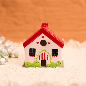 Può includere: Una statuetta a forma di casa in ceramica con tetto rosso e pareti bianche. La casa ha finestre quadrate nere, un'apertura rotonda e una porta ad arco verde e rossa. Piccoli cuori rossi decorano il tetto e le pareti. Dettagli verdi simili all'erba alla base.