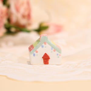 Op de afbeelding: Een miniatuur keramisch huisje met een witte basis, een rode deur en een groen dak. Het dak is versierd met kleurrijke accenten. Het huisje is een charmant decoratief item.