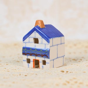 May include: A miniature ceramic house with a blue roof, white walls, and an orange chimney. The house has small square windows and decorative blue and green trim. The overall design is reminiscent of a Mediterranean style.