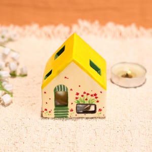 Puede incluir: Un portavelas de cerámica con forma de casa, con tejado amarillo y paredes color crema. La casa tiene marcos de ventanas y puertas verdes, con detalles de flores rojas. Una vela de té encendida está cerca, creando un ambiente cálido.