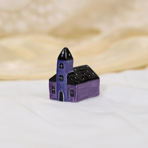 Może przedstawiać: Miniaturowy ceramiczny kościół z fioletowym korpusem i czarnym dachem i wieżą. Wieża jest w ciemniejszym odcieniu fioletu. Kościół ma małe okna w kształcie łuku i krzyż na wieży. Jest to przedmiot dekoracyjny.