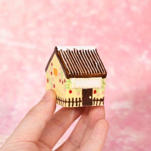 May include: A miniature ceramic house with a yellow facade, brown roof, and white accents. The house features a small door, window, and a decorative fence. Floral and fruit designs adorn the walls. The house is held in a hand against a pink background.