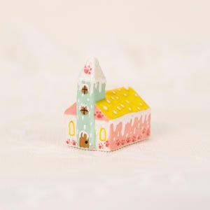 May include: A miniature church figurine with a white steeple, a mint green tower, and a yellow roof. The church is painted with pink and white accents, and paw prints decorate the base and steeple.
