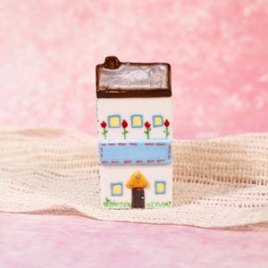 May include: A whimsical, rectangular ceramic house figurine. The house features a brown roof, white walls with red flower accents, and a blue banner. A yellow and orange triangle door is at the base. The figurine is on a white, textured surface.