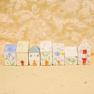 May include: A collection of seven ceramic houses, each uniquely decorated with floral and leaf patterns. The houses feature different colored roofs, doors, and windows, with a whimsical, handcrafted aesthetic. The houses are arranged in a row.