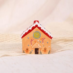 May include: A miniature gingerbread house with orange walls, a red roof, and white trim. The house features a dark brown door, candy cane accents, and gingerbread man figures. A blue and yellow circular design is centered on the front.