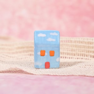 May include: A small, rectangular ceramic house figurine. The house is painted light blue with white cloud accents. It has two orange square windows and a red door. The background is a soft pink.