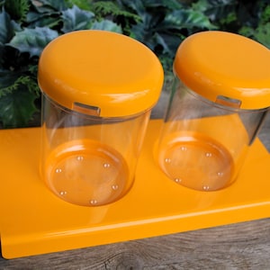 Peut inclure: Deux récipients cylindriques transparents avec des couvercles orange sont posés sur un plateau rectangulaire orange. Les récipients ont de petites indentations circulaires au fond. Les couvercles ont un petit fermoir rectangulaire.