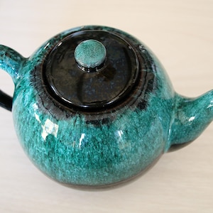 Peut inclure: Une théière en céramique avec un corps arrondi, une anse incurvée et un bec verseur. La théière est émaillée dans des tons de vert d'eau et de noir, avec un couvercle et un bouton assortis. C'est un article de cuisine.