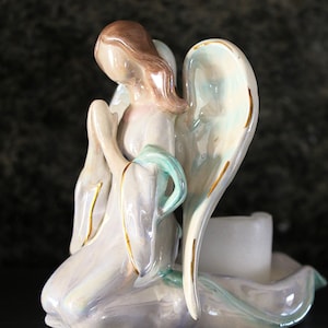 May include: A ceramic angel figurine kneeling with folded hands and large wings. The angel is painted in soft pastel colours, with gold accents on the wings and clothing. A white candle holder is integrated into the base.