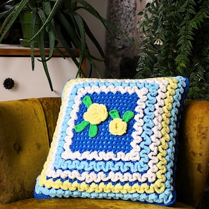 Peut inclure: Un coussin carré au crochet avec un motif floral. Le coussin présente un centre bleu avec des appliqués de fleurs jaunes et vertes. La bordure comporte des sections blanches, bleues et jaunes. Le coussin est sur une chaise en velours doré.