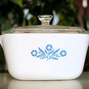 May include: White casserole dish with a clear glass lid. The dish has a blue floral design. It has two handles and is likely made of ceramic. The dish is suitable for cooking.