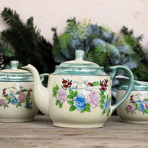 Peut inclure: Service à thé crème composé d'une théière, d'un sucrier et d'une crémière. Chaque pièce présente un motif floral avec des accents bleus, roses et verts, et une scène de paysage. Les bords et les poignées sont vert clair.