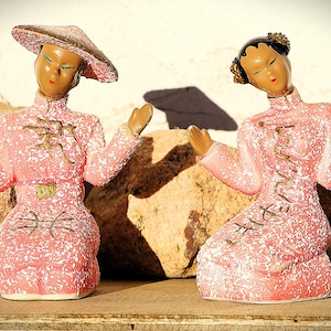 Peut inclure: Deux figurines en céramique de femmes asiatiques en robes roses à mouchetures blanches. L'une porte un chapeau, l'autre a les cheveux relevés. Les deux ont les bras levés et sont à genoux. Des accents dorés ornent les robes.