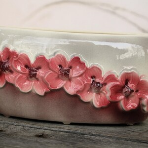 May include: Oval ceramic planter in shades of grey and mauve, decorated with a row of pink floral embellishments. The flowers have detailed petals and dark centres, adding elegance. The planter is approximately 25 cm long.