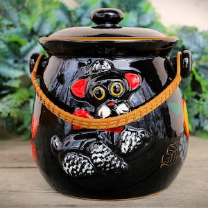 Peut inclure: Pot à biscuits en céramique noire avec couvercle et anse tressée. Le pot présente un motif fantaisiste d'un chien de dessin animé avec des oreilles rouges et un museau blanc. Des accents décoratifs de fruits et de fleurs sont également présents.