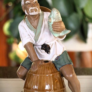 Peut inclure: Figurine en céramique d'un pêcheur âgé tenant un poisson. La figurine porte une tunique blanche, un short vert et est assise sur un tonneau marron. La figurine a un visage détaillé et est peinte dans des tons terre.