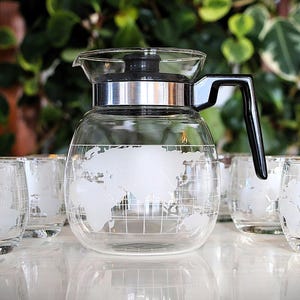 May include: A clear glass carafe and matching glasses set. The carafe has a black handle and a silver band. Each piece features a white world map design. The set is ideal for serving beverages.