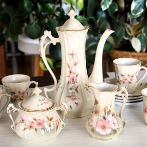 May include: A cream-coloured porcelain tea set with pink floral patterns. The set includes a tall teapot, sugar bowl, creamer, and several teacups and saucers. Gold trim accents the edges of each piece, creating an elegant aesthetic.