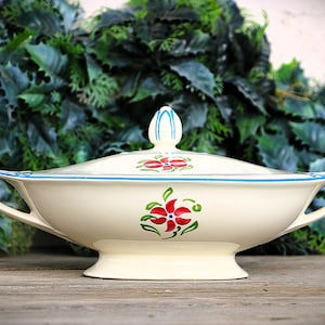 May include: Cream-coloured ceramic serving dish with a lid, featuring red and green floral designs. The dish has a blue trim and handles. The background is a blurred arrangement of green leaves.