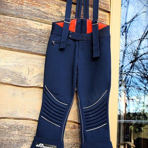 May include: Navy blue ski trousers with red detailing and braces. The trousers have white piping, quilted knee sections, and the "Schneider" logo on the lower leg. A vintage ski apparel item.