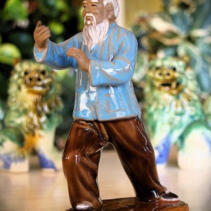 Peut inclure: Figurine en céramique d'un vieil homme en veste bleue et pantalon marron, en position d'arts martiaux. La figurine a une barbe blanche et se tient sur une base marron. Deux statues de lions décoratives sont en arrière-plan.