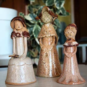 May include: Three ceramic figurines in a row, each with a unique design. The figurines are in shades of brown and beige, with detailed facial features and clothing. The figurines are approximately 15 cm tall.
