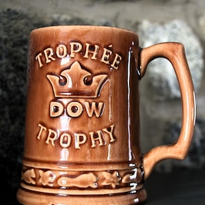 Peut inclure: Chope à bière en céramique marron avec les mots "TROPHEE DOW TROPHY" en lettres en relief. La chope présente un motif de couronne et une anse. La chope est probablement vintage et a une finition brillante.