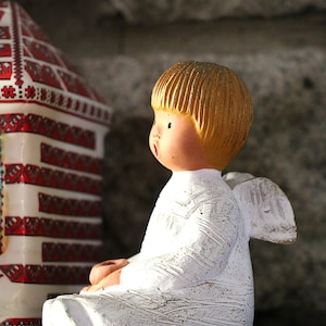 Peut inclure: Figurine d'ange en céramique avec une tête jaune doré et une robe blanche. L'ange a de petites ailes blanches et est assis, les mains jointes. Une structure décorative en céramique avec des motifs rouges et blancs est en arrière-plan.
