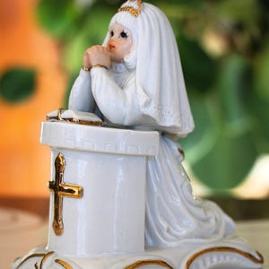May include: A white porcelain figurine of a girl kneeling in prayer. The figurine wears a white dress and veil, with gold accents on the cross, base, and details. The girl is kneeling in front of a white pedestal.