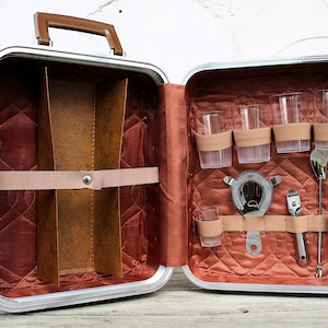 Peut inclure: Un ensemble de bar portable dans une mallette rigide argentée avec une poignée marron. L'intérieur est doublé de tissu matelassé rouille et comporte des sangles élastiques retenant quatre verres cannelés, une passoire, un doseur et une cuillère.