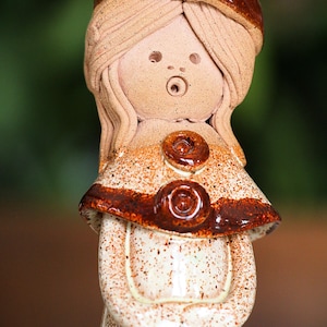 May include: A handmade ceramic figurine of a girl with long hair and a surprised expression. The figurine has a brown and tan colour scheme with a speckled pattern. The girl is wearing a dress and a decorative shawl with two large buttons.