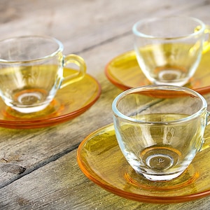 May include: Three clear glass teacups with yellow handles and matching saucers. The cups and saucers are arranged on a weathered wooden surface, creating a warm aesthetic. The transparent design allows light to pass through, highlighting the golden hue.