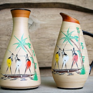 Peut inclure: Un vase en céramique et une carafe assortie avec une finition imitation bois beige clair. Les deux pièces présentent des illustrations peintes à la main de trois personnages dans une barque sous des palmiers, vêtus de jupes jaune, blanche et rouge.