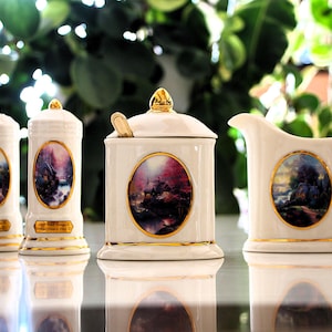 Peut inclure: Un ensemble de quatre articles de cuisine en céramique blanche avec des bordures dorées. L'ensemble comprend deux salières et poivrières, une sucrière avec une cuillère et une crémière. Chaque pièce présente une peinture ovale avec un paysage.