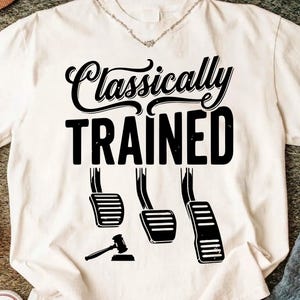 Może przedstawiać: Biała koszulka z czarnym napisem "Classically TRAINED", z ilustracją pedałów samochodowych i młotka. Koszulka ma okrągły dekolt i srebrny naszyjnik.