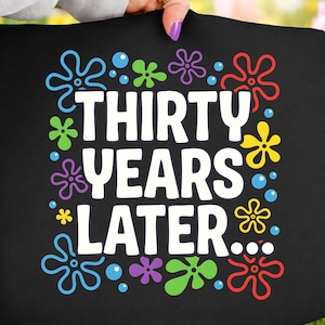 May include: Black tote bag with the text "THIRTY YEARS LATER..." in white, surrounded by colorful flower and bubble graphics. The bag is held up by a person with a gold ring and purple nail polish.