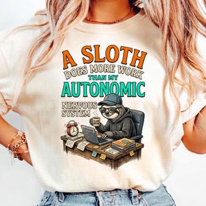 Könnte beinhalten: Cremefarbenes T-Shirt mit dem Text "A Sloth Does More Work Than My Autonomic Nervous System." Die Grafik zeigt ein Cartoon-Faultier mit Brille und Hut, das an einem Schreibtisch mit Laptop, Kaffeetasse und Wecker arbeitet.