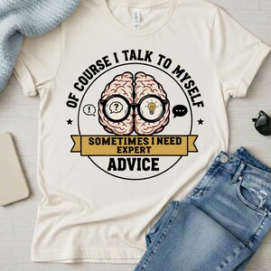 May include: Cream-colored t-shirt with a graphic of a brain wearing glasses, with the text "OF COURSE I TALK TO MYSELF" and "SOMETIMES I NEED EXPERT ADVICE". The shirt is paired with blue jeans and a light blue sweater.