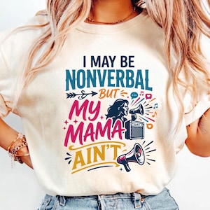 Op de afbeelding: Crèmekleurig T-shirt met de tekst "I MAY BE NONVERBAL BUT MY MAMA AIN'T" in teal, roze en geel. De afbeelding bevat een vrouw met een megafoon, een versterker en muzieknoten.