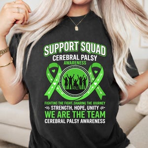 May include: Black t-shirt with a green and white design promoting Cerebral Palsy awareness. The design includes the words "Support Squad," green ribbons, and the phrases "Fighting the Fight, Sharing the Journey" and "We Are The Team."
