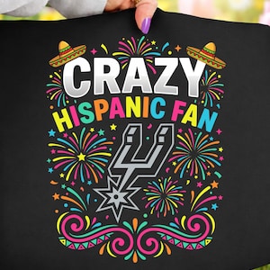 May include: Black t-shirt with the words "CRAZY HISPANIC FAN" in white and yellow, featuring a team logo and colourful fireworks and sombreros. The design is vibrant and festive.
