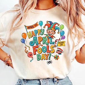 Könnte beinhalten: Cremefarbenes T-Shirt mit einem farbenfrohen "Happy April Fools' Day!"-Motiv. Das Design umfasst einen Narren, Luftballons und die Wörter "April 1st" und "Prank On!" in einer verspielten Schriftart. Ein lustiges, festliches Design.
